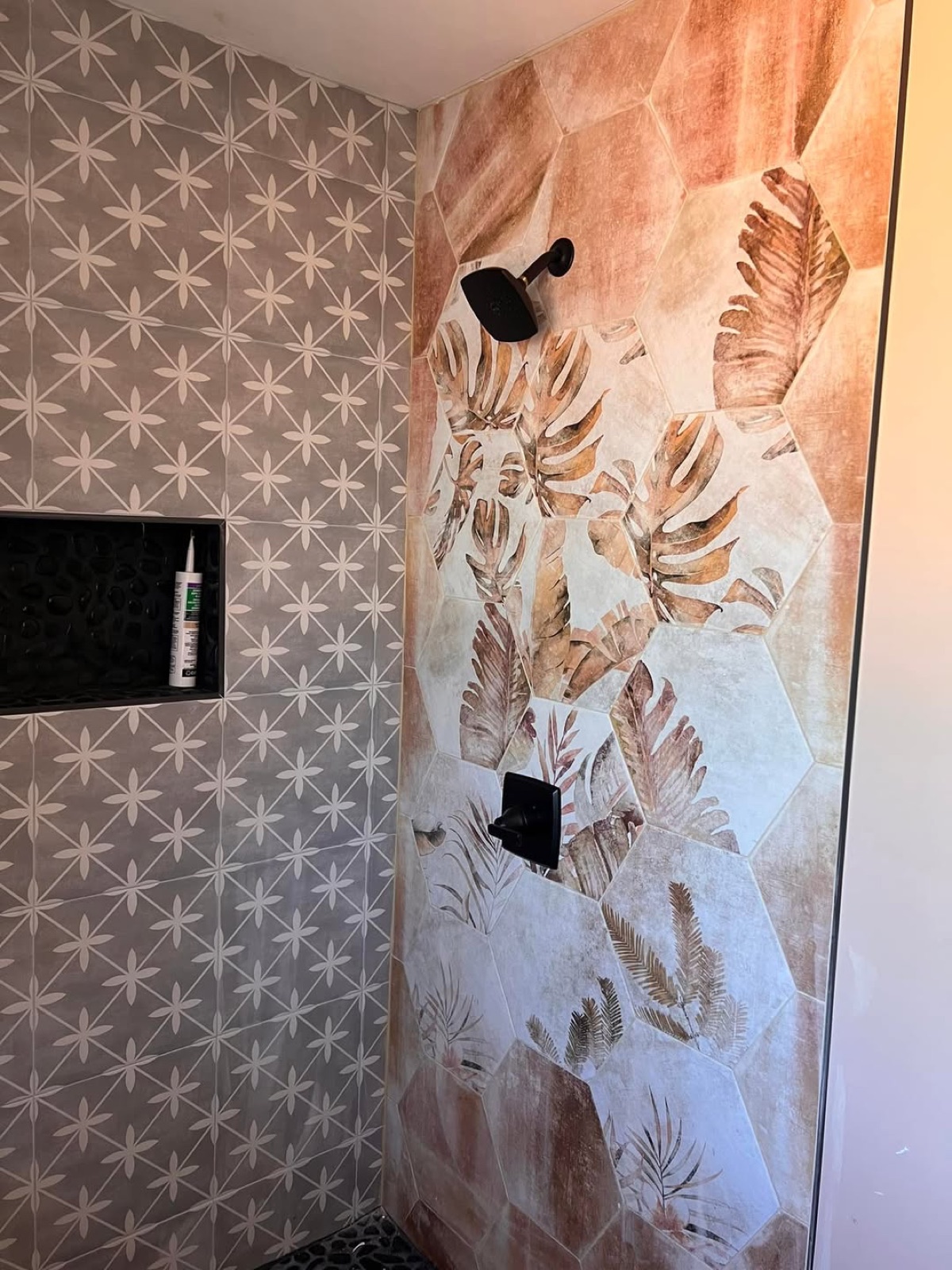 Custom walk-in shower with patterned gray floral tile beside a botanical leaf mural tile wall during installation