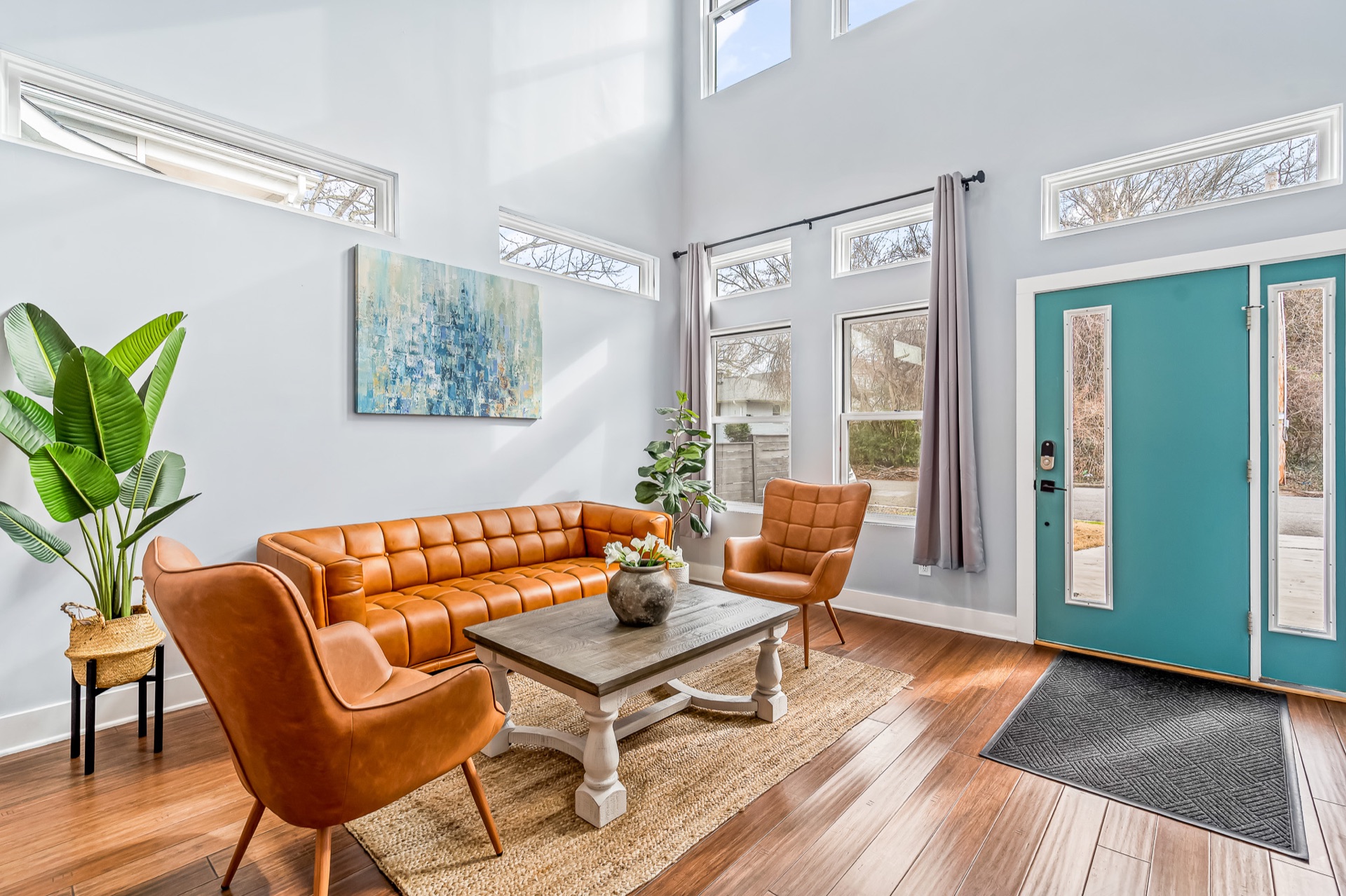 Bright two-story living room with tan leather furniture, a teal entry door, and warm hardwood flooring