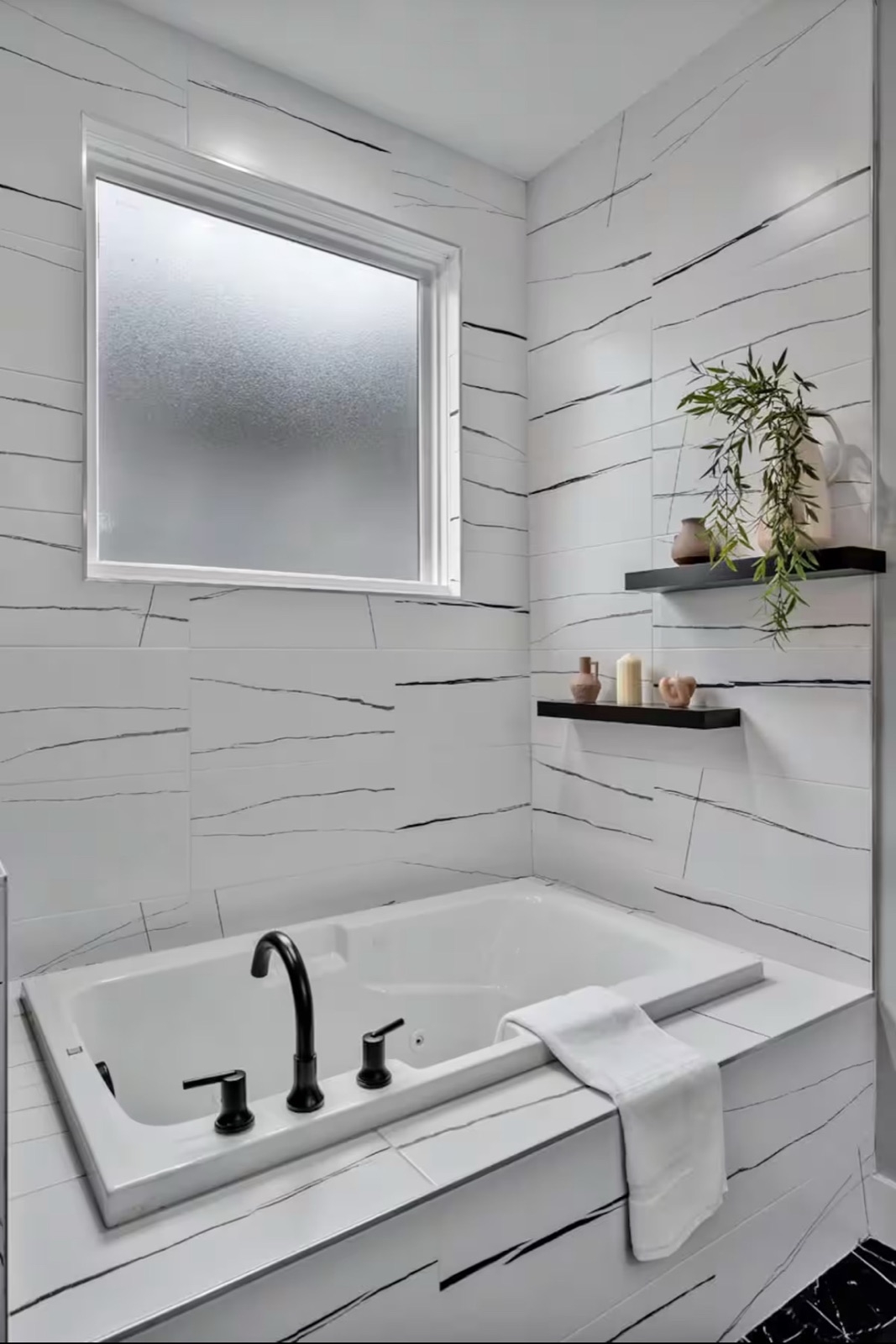 Built-in soaking tub with white marble-veined tile surround, matte black fixtures, frosted window, and floating shelves with plants