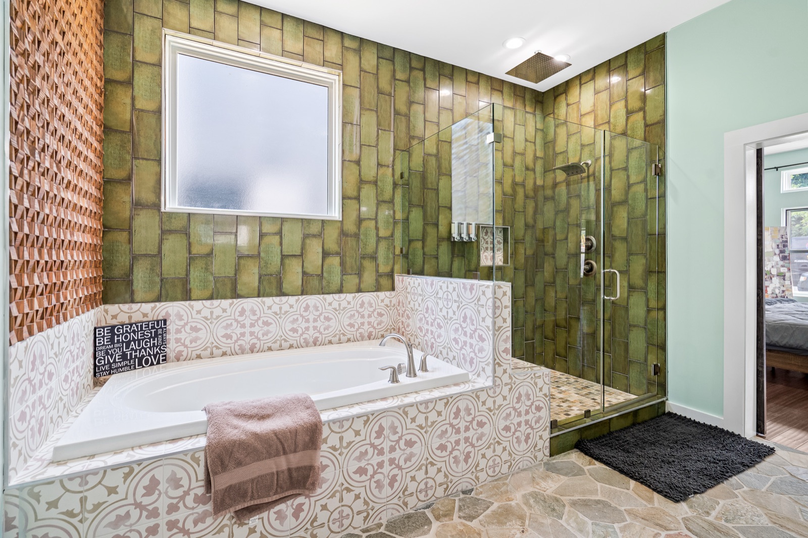 Soaking tub with patterned tile surround beside a frameless glass shower in a green-tiled renovated bathroom