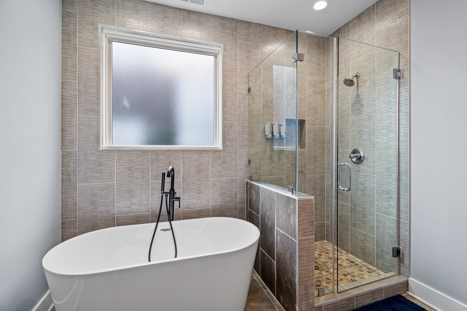 Freestanding white soaking tub with matte black filler beside a frameless glass walk-in shower with wood-look tile