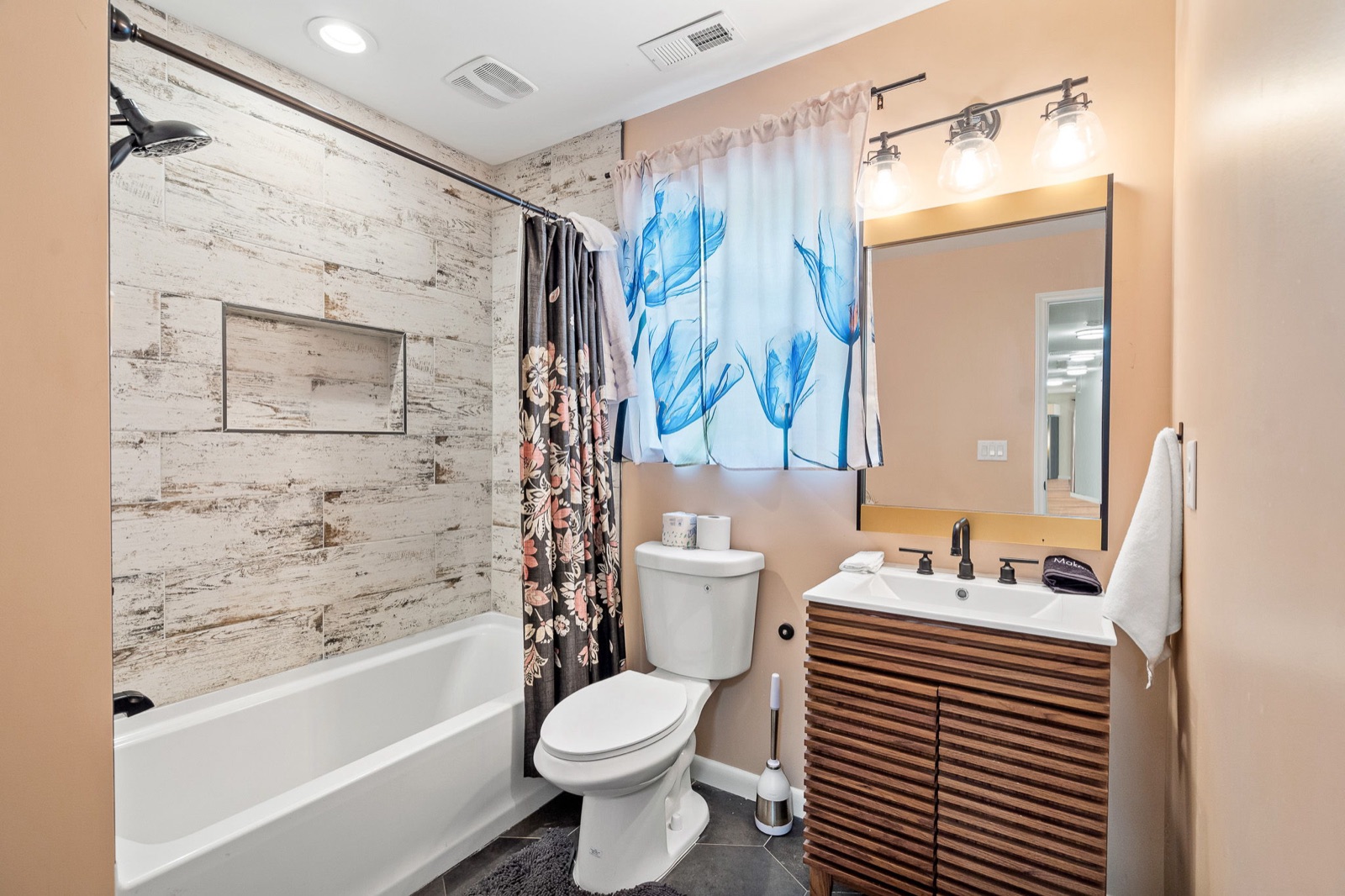 Updated full bathroom with distressed wood-look tile tub surround, modern slat-front vanity, framed mirror, and toilet