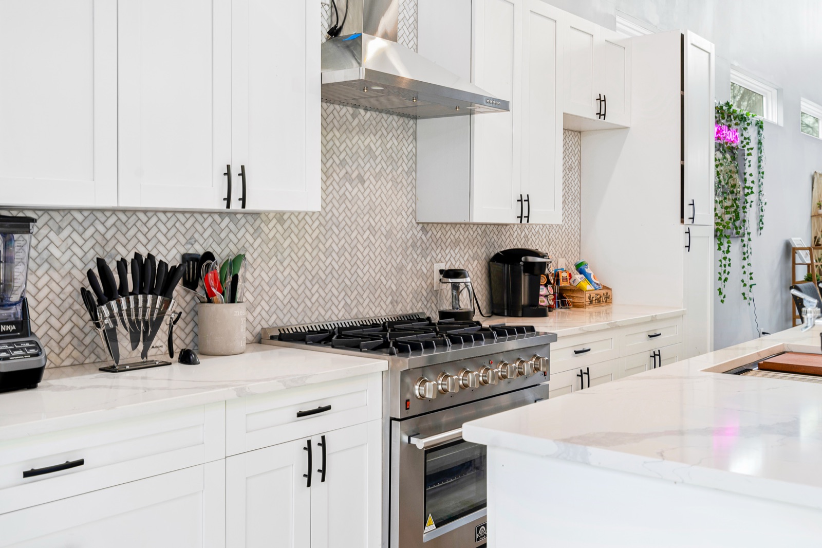 White shaker kitchen with stainless gas range, herringbone marble backsplash, and waterfall quartz island