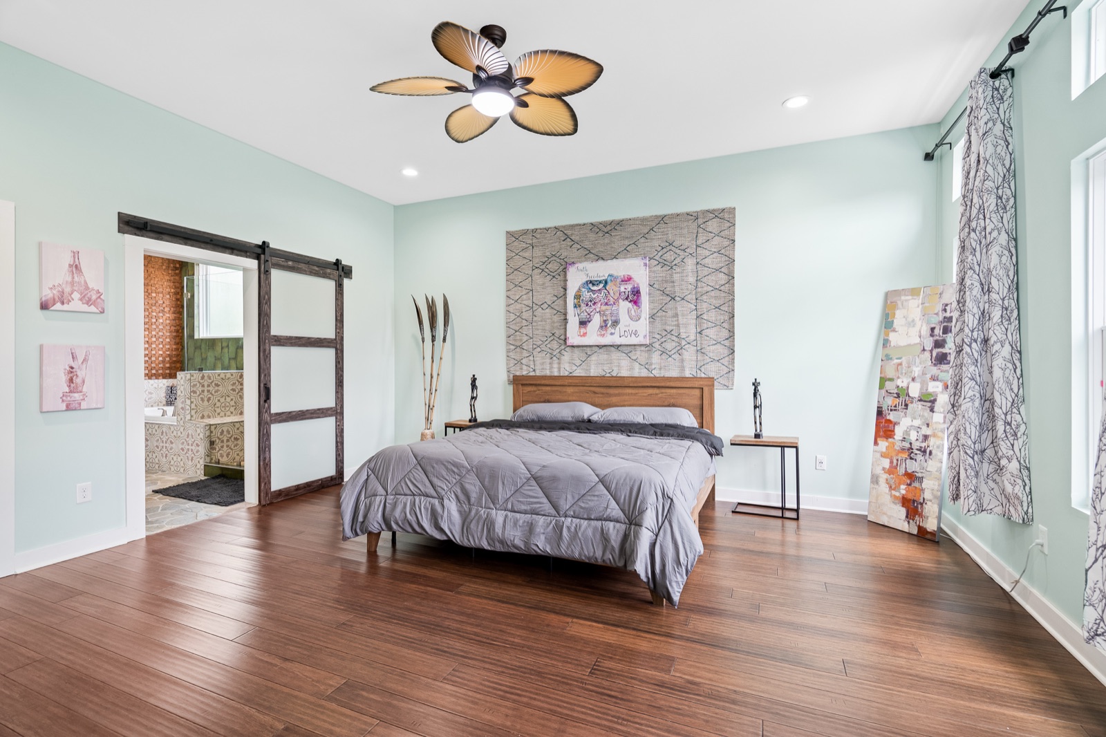 Large bedroom with mint walls, hardwood floors, gray-bedded king bed, and a barn door leading to an en-suite bathroom