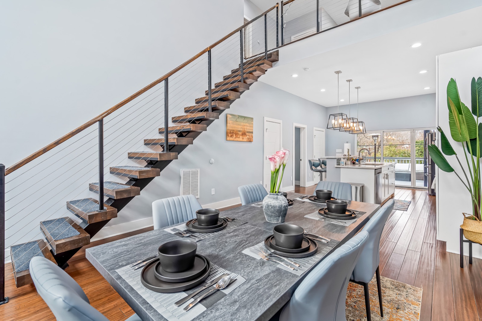 Staged dining table set for four next to a modern floating wood staircase with cable railings and an open white kitchen beyond
