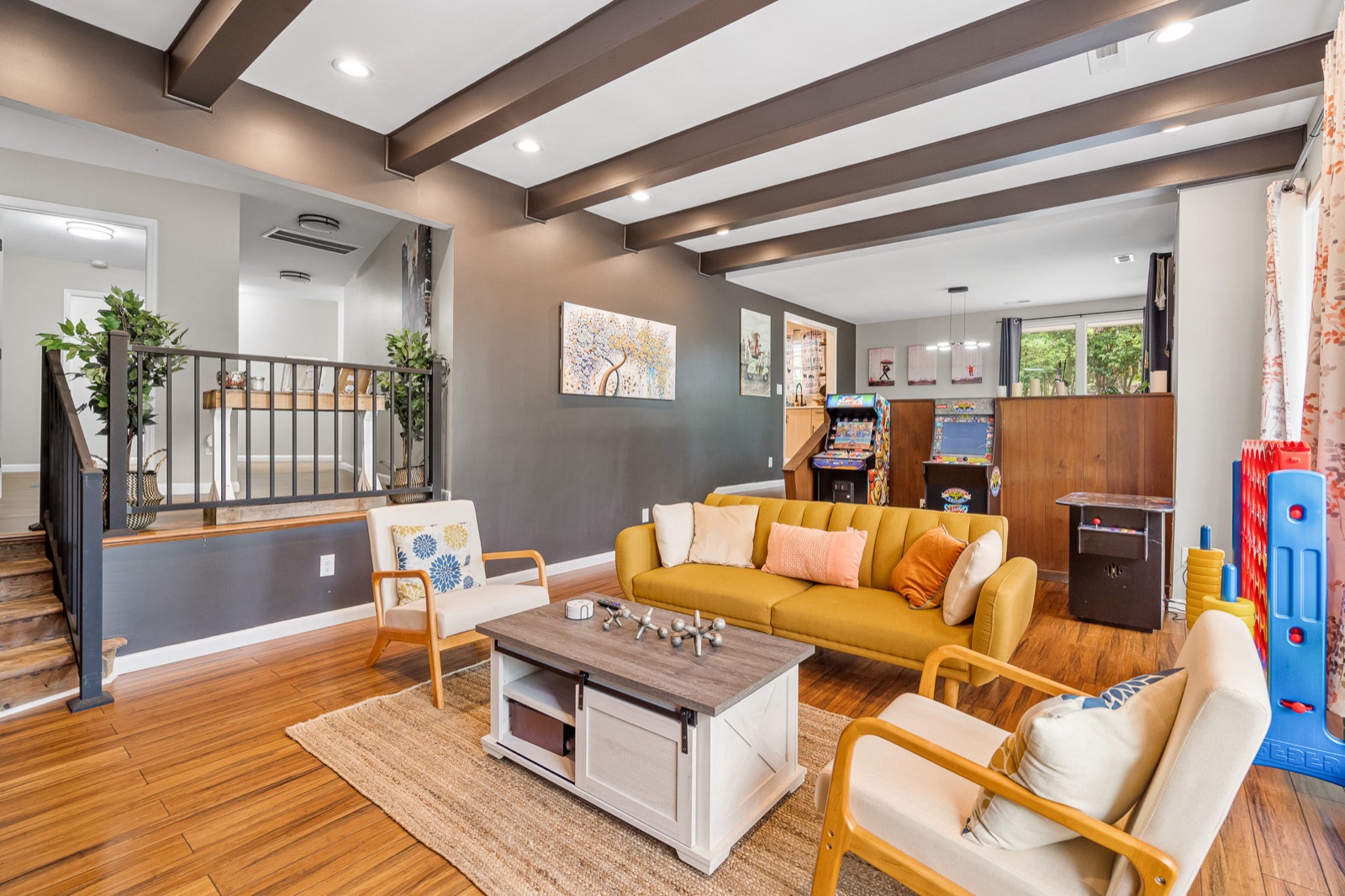 Open-concept living and game room with exposed beams, mustard seating, hardwood floors, and a staircase railing