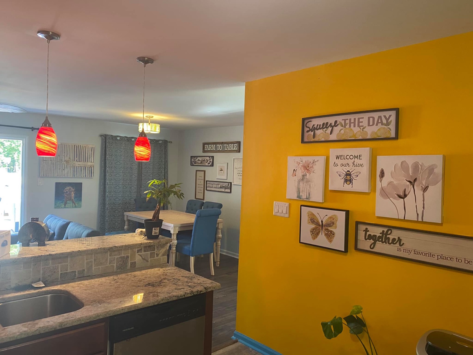 Open kitchen and dining space with granite peninsula, stainless sink, and a vibrant yellow accent wall with framed botanical and bee art