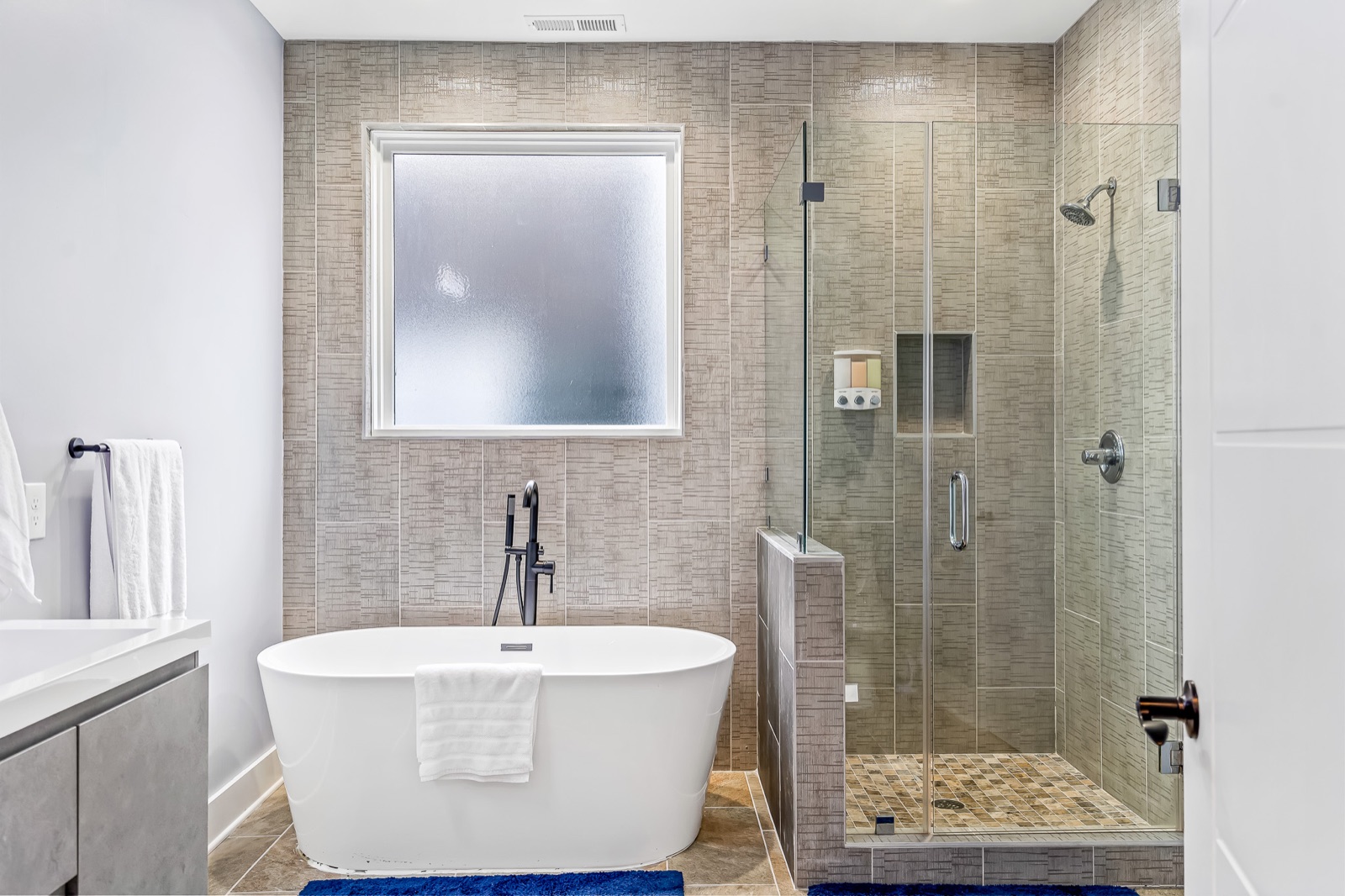 Renovated primary bathroom with freestanding soaking tub, frameless glass walk-in shower, vertical tile, and matte-black fixtures